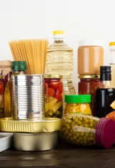 Various pantry foods, such as fruits, vegetables, beans, crackers, and pasta, packaged in glass jars, aluminum cans and tins, plastic bottles and other plastic packaging.